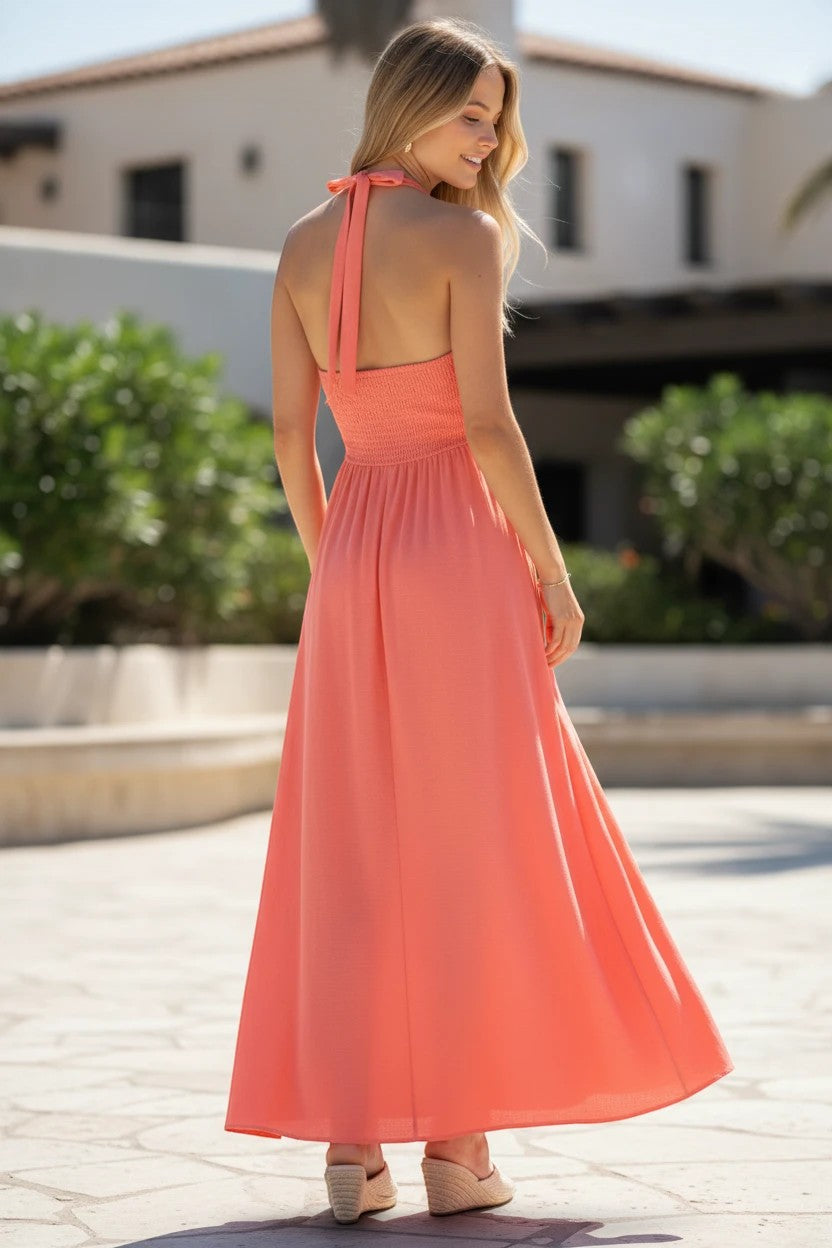 Robe longue Valerelle corail à col licou et dos nu, portée par une femme dans un cadre estival ensoleillé.