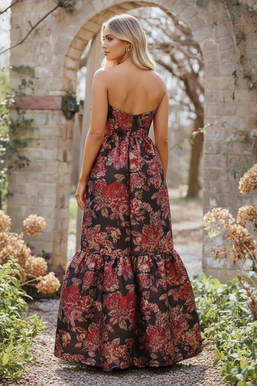 Robe de soirée longue Valerelle à imprimé floral rouge et noir, coupe bustier fluide dans un jardin en pierre.