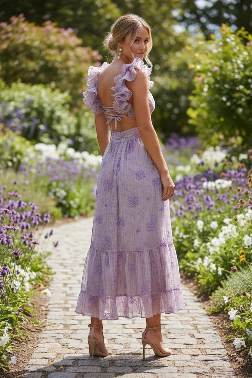 Robe de soirée longue Valerelle lilas à volants et dos nu, portée par une femme dans un jardin fleuri.