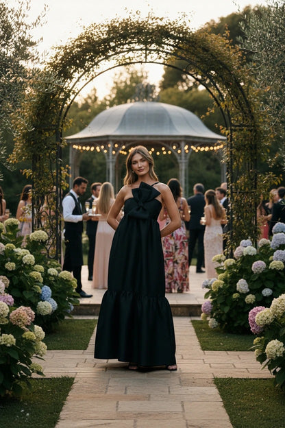 Robe de cocktail longue noire Valerelle avec nœud décoratif à la taille, portée par une femme dans un jardin chic.