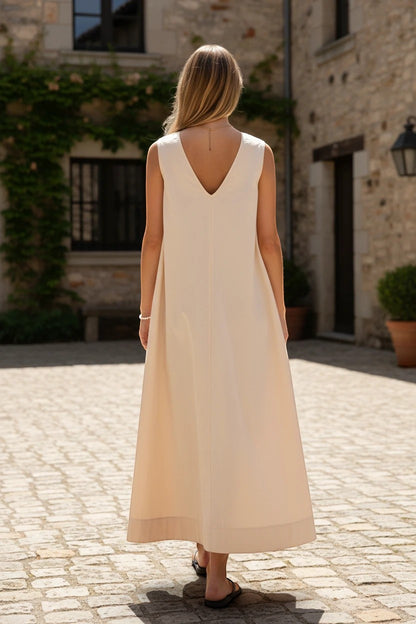 Robe de demoiselle d'honneur Valerelle longue beige avoine fluide avec dos nu en V, vue de dos dans une cour pavée.