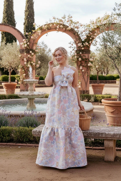 Robe longue Valerelle asymétrique à imprimé floral avec nœud sur l'épaule, portée dans un jardin ensoleillé.