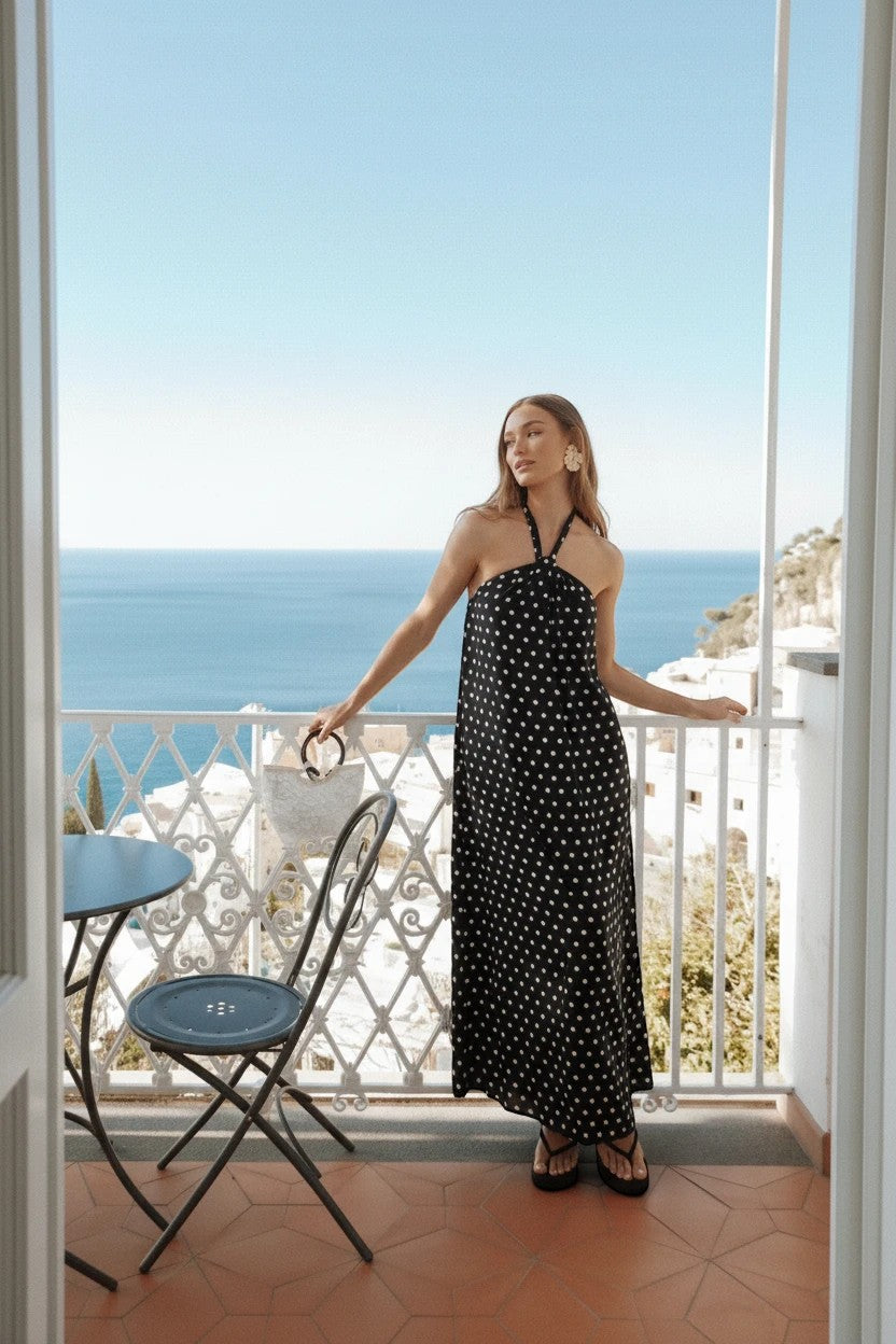 Robe longue Valerelle noire à pois blancs, coupe fluide avec encolure dos nu, portée sur une terrasse ensoleillée.