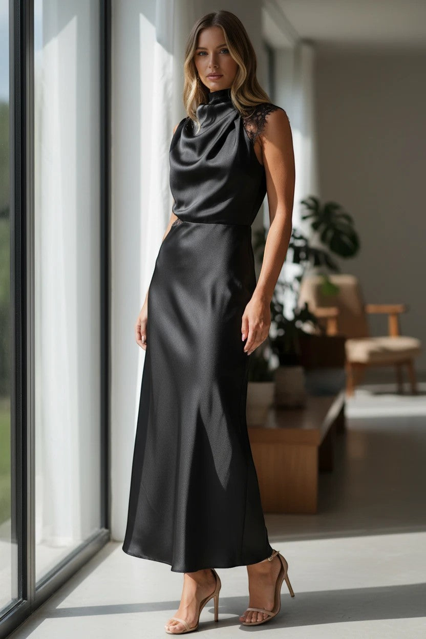 Robe longue Valerelle noire en satin et dentelle avec col licou drapé portée par une femme près d'une fenêtre