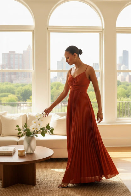 Robe longue plissée orange Valerelle à fines bretelles portée par une femme dans un salon lumineux avec vue urbaine