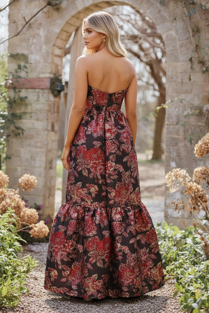 Robe de soirée longue Valerelle à imprimé floral rouge et noir, coupe bustier fluide dans un jardin en pierre.